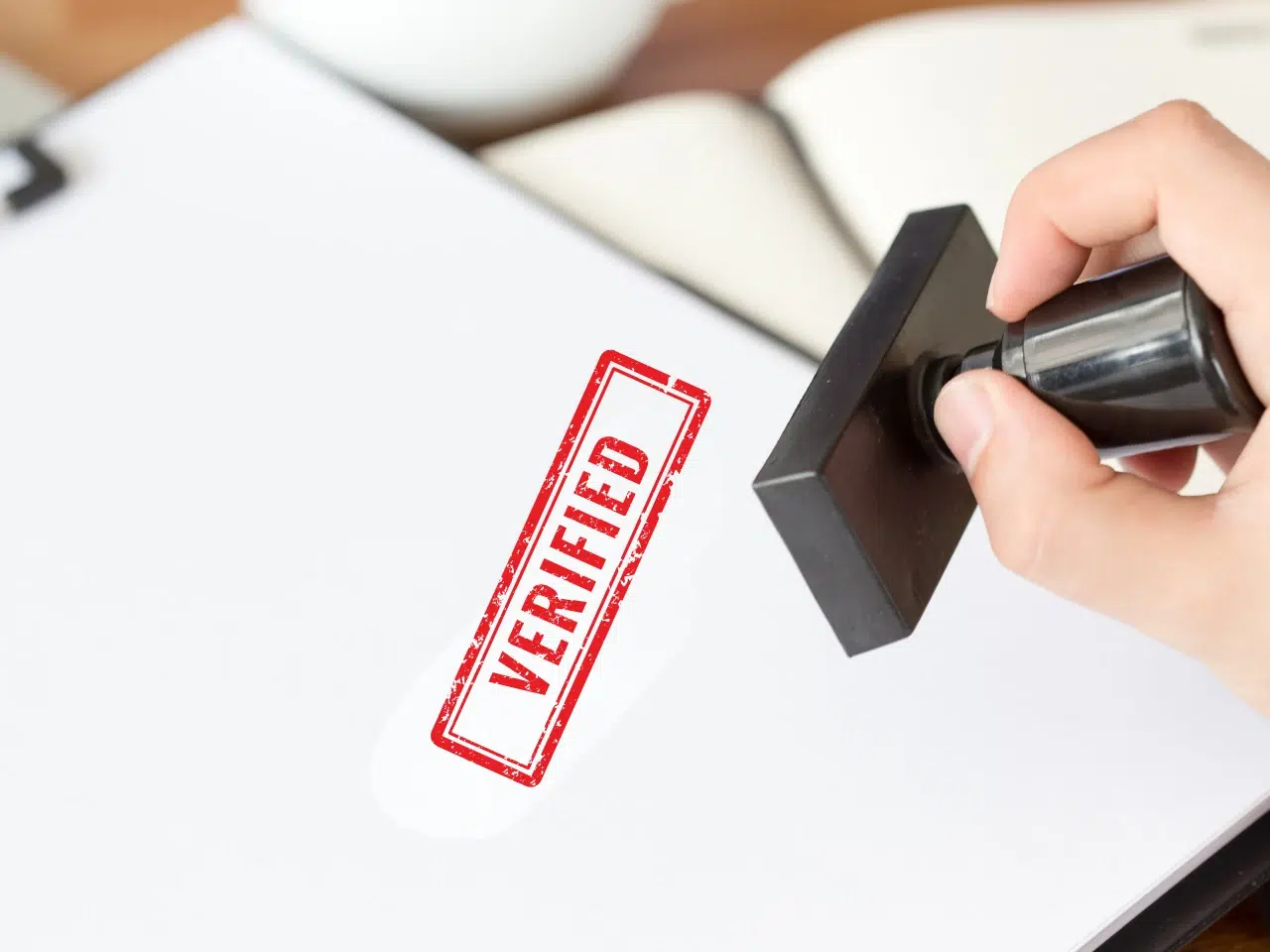 A hand is holding a stamp and stamping a red VERIFIED mark on a white sheet of paper, with a blurred notebook in the background.