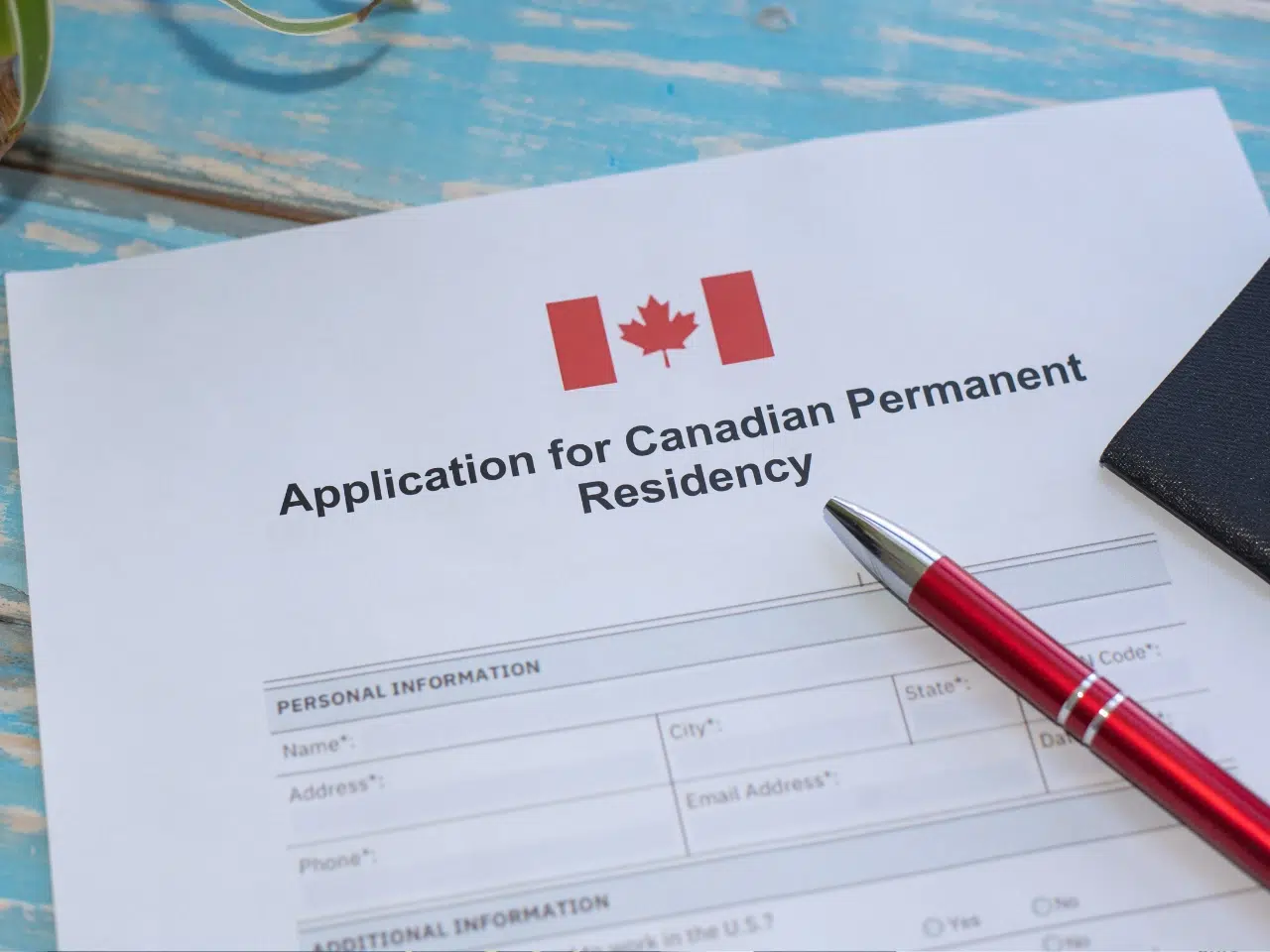 A Canadian permanent residency application form with a red pen placed on top, set on a light blue wooden surface. The form features the Canadian flag and fields for personal information.