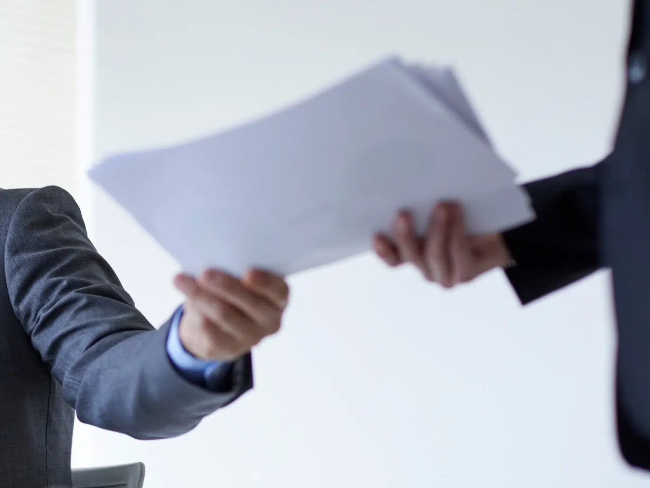 A person in a suit hands a stack of papers to another person, both wearing business attire. The background is out of focus and bright, suggesting an office environment.