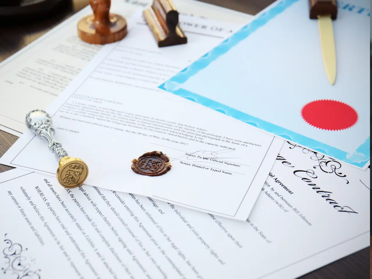 Several official documents and certificates are spread out on a table, featuring a wax seal, a sealing stamp, a stamp pad, and a letter opener. One document has a red embossed seal.