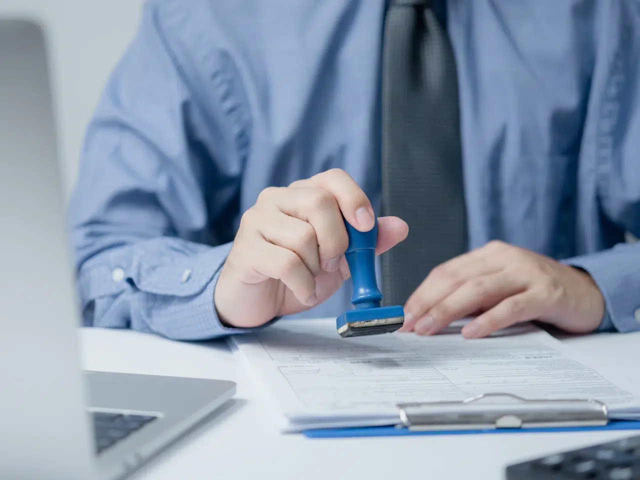 A person in a blue shirt and tie holds a blue stamp, pressing it onto a document on a desk. A laptop and clipboard with papers are also visible on the desk.
