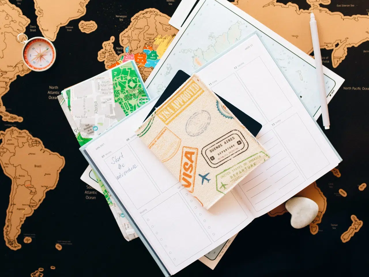 A flat lay of travel essentials, including a passport cover, open planner, maps, a compass, a pen, and a small white stone, arranged on a world map background.