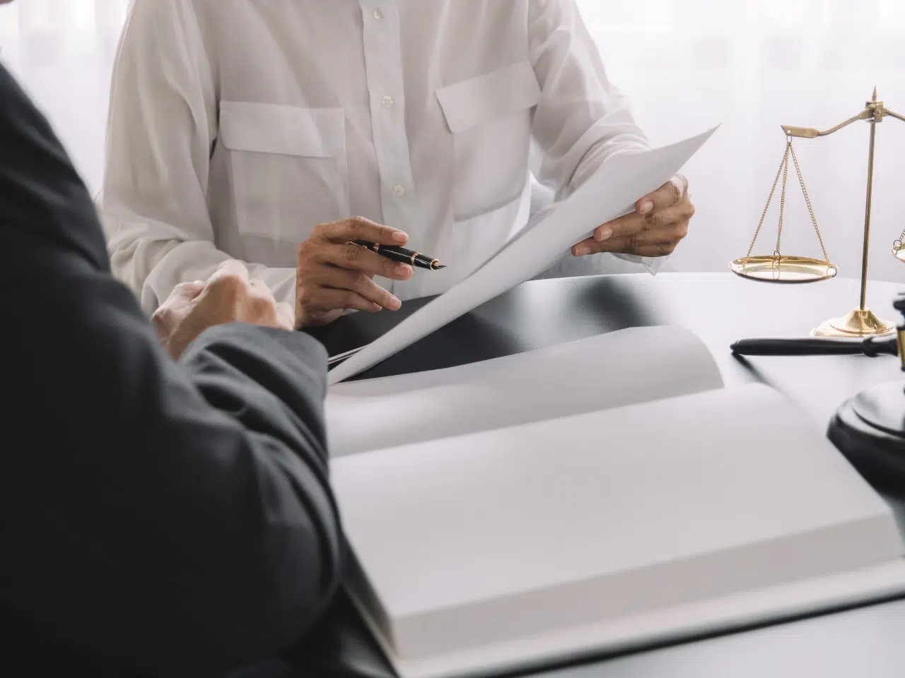 Two people sit at a desk reviewing documents. One person holds papers and a pen, while the other listens. A scale of justice and a gavel are on the table, suggesting a legal or consultation setting.