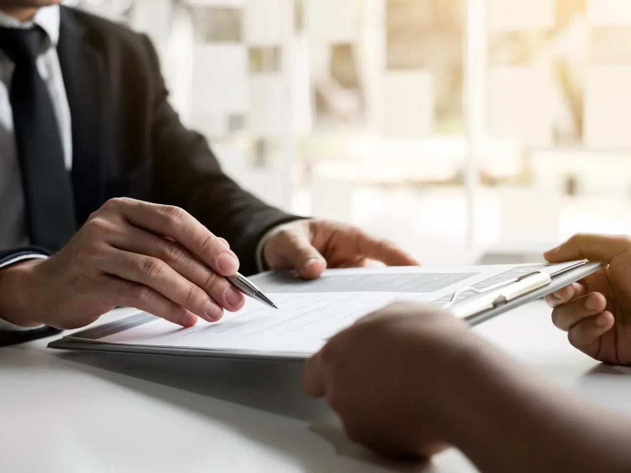 Two people in formal attire sit at a desk; one holds a clipboard with documents while the other points at the papers with a pen, suggesting a discussion or review of business or legal paperwork.