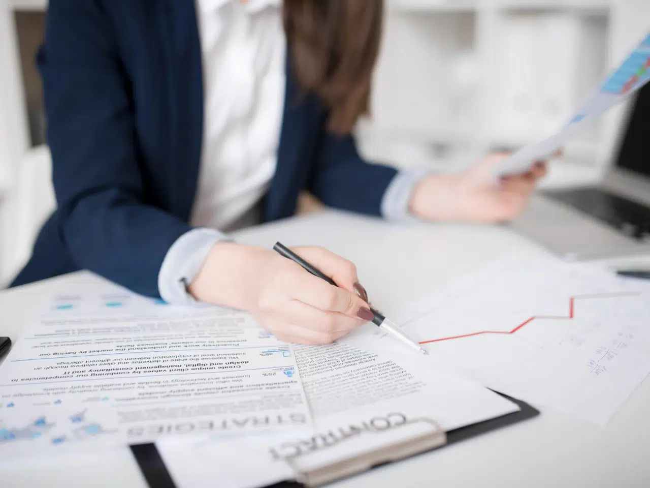 A person in business attire sits at a desk, reviewing documents and charts. One hand holds a paper, while the other writes on a contract form with graphs and data scattered around. The background is an office setting.