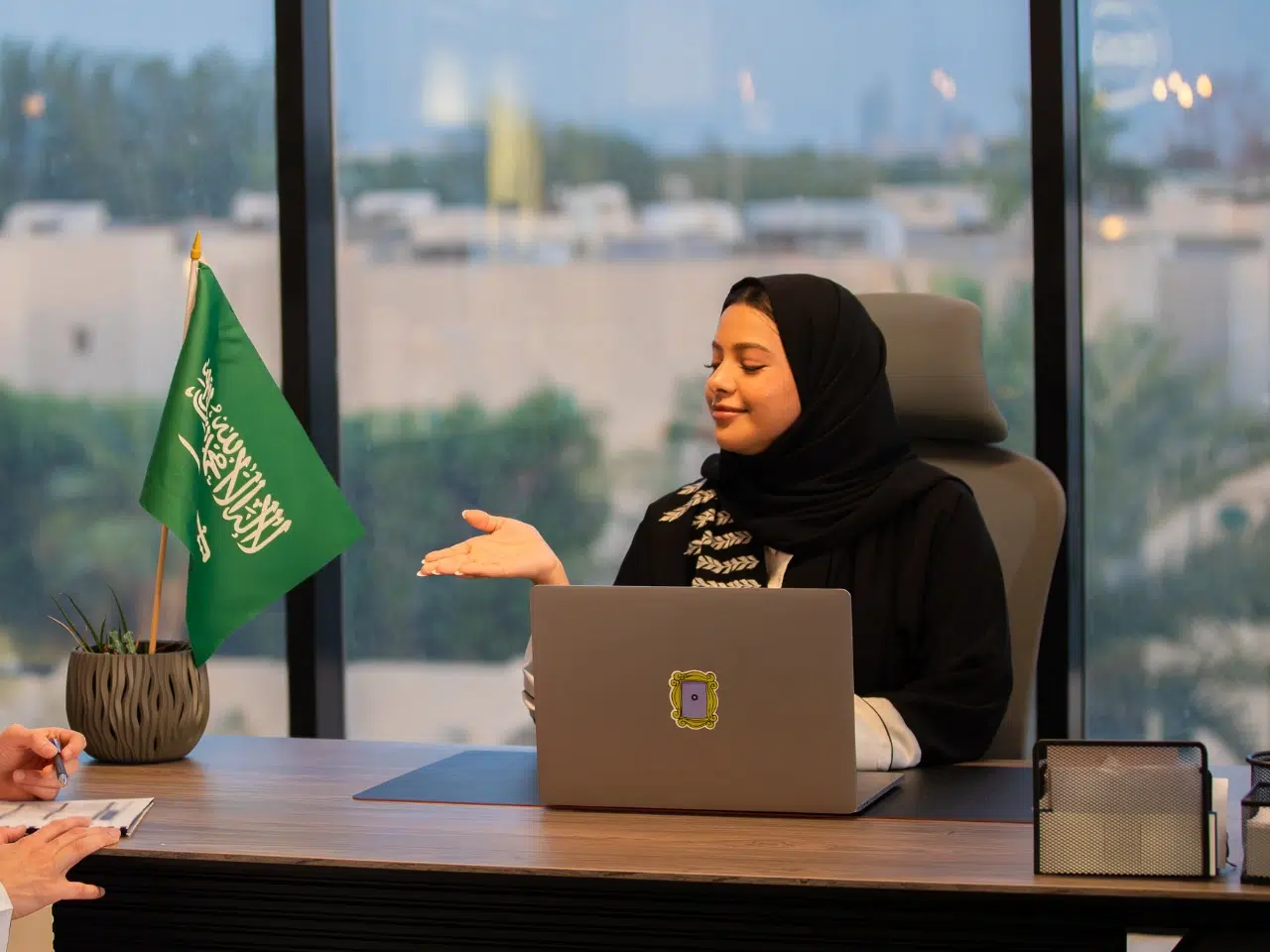 A woman wearing a black hijab sits at an office desk with a laptop, gesturing as she speaks. A Saudi Arabian flag and a potted plant are on the desk, and a large window shows a blurred outdoor view.