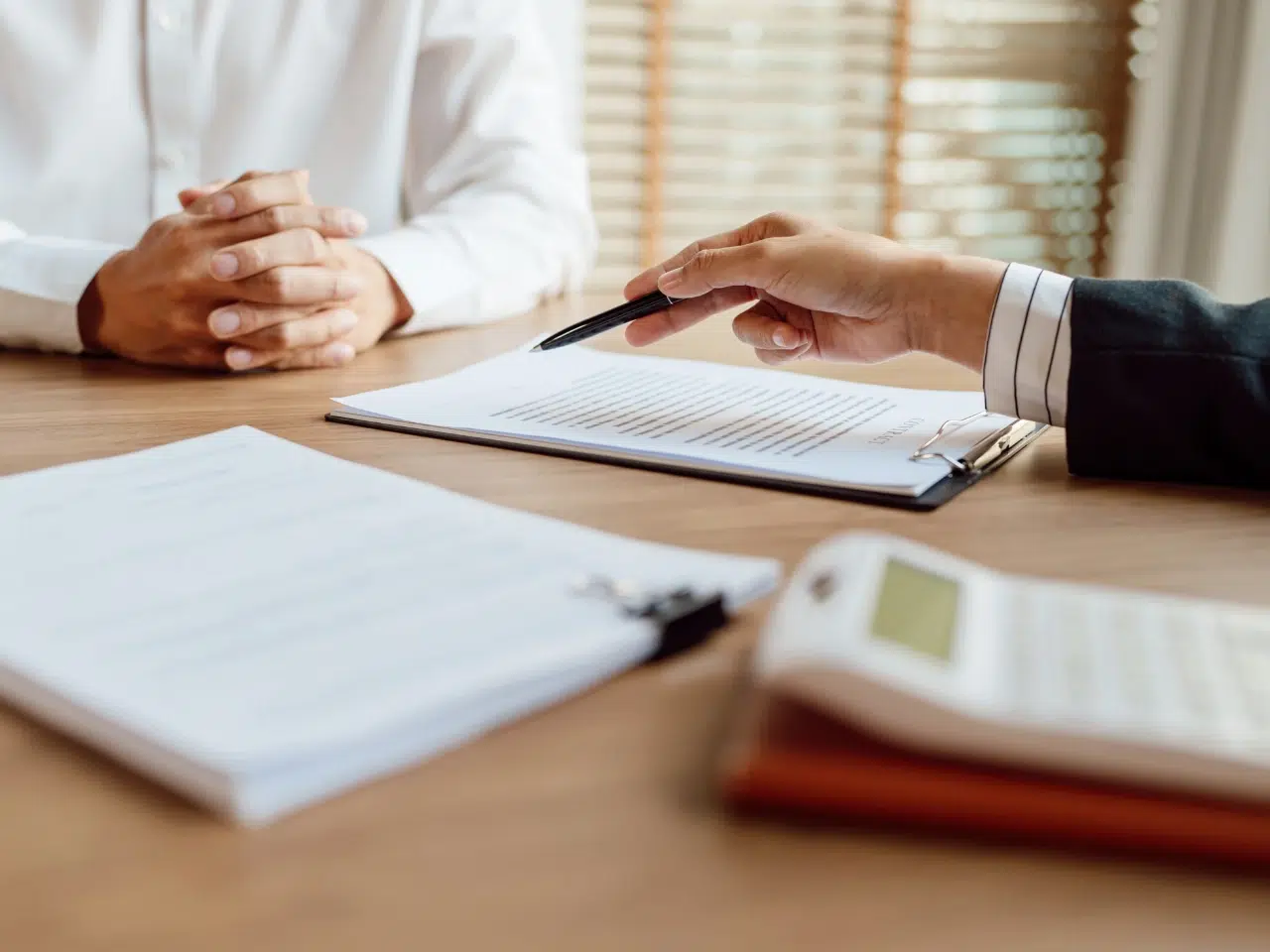 Two people sit at a desk with documents and a calculator. One person points to a clipboard with papers while the other has hands folded, suggesting a discussion or agreement review.