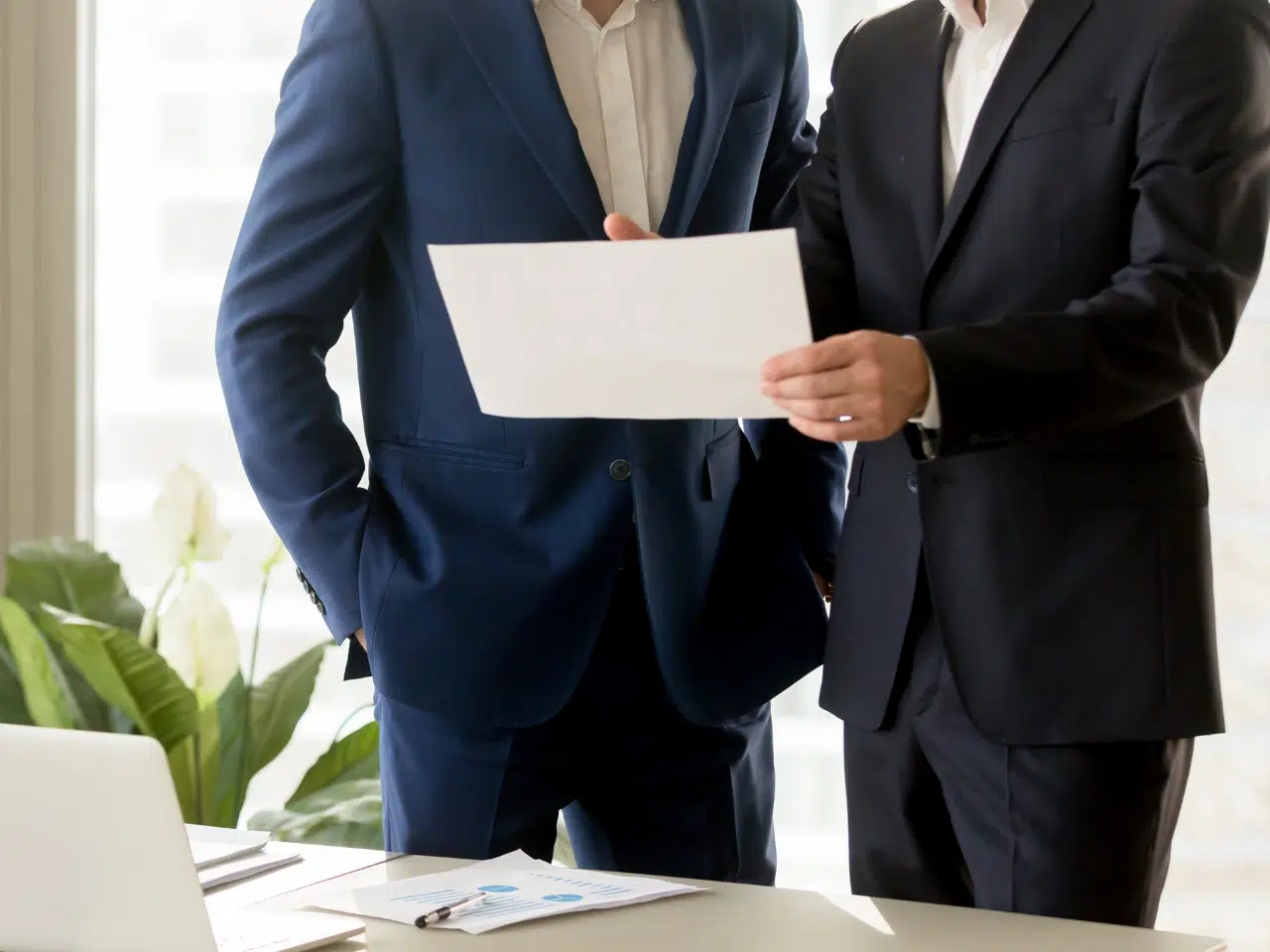 Two men in suits stand by a desk, holding and reviewing a sheet of paper together. Office supplies and a laptop are on the desk, with plants and large windows in the background.
