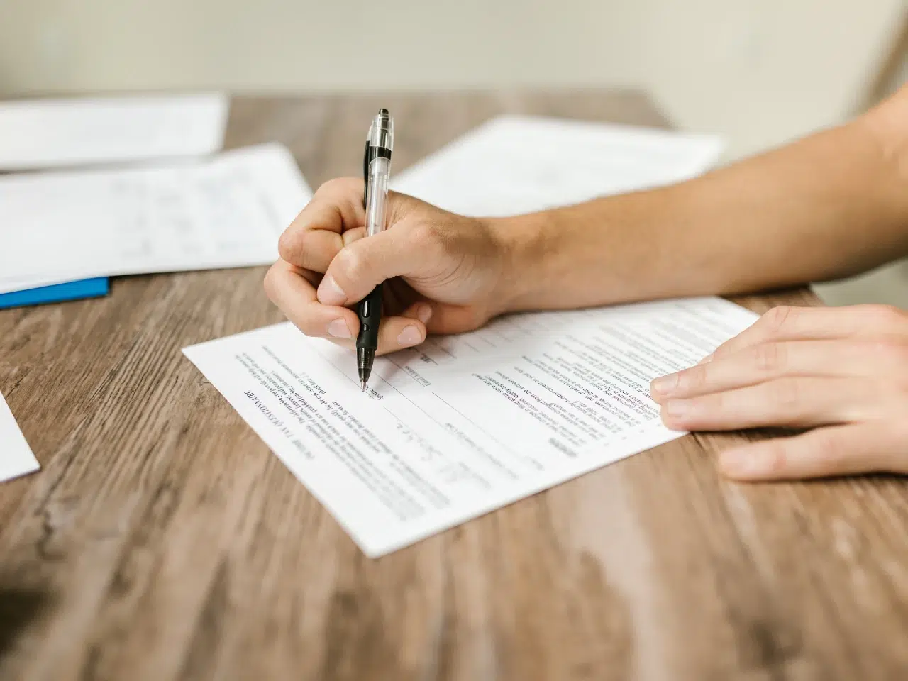 A person holding a pen fills out a form on a wooden table, with additional papers in the background.