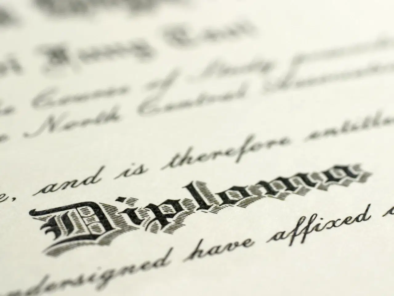 Close-up of the word Diploma written in ornate, black Gothic-style lettering on a formal certificate, with blurred cursive text visible in the background.