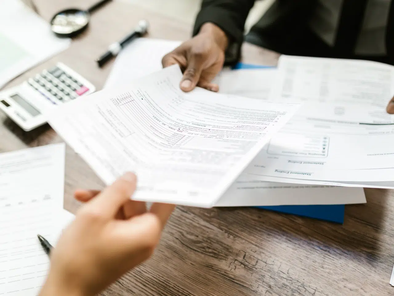 Two people exchange paperwork at a wooden desk with documents, a calculator, a pen, and a magnifying glass, suggesting a business or financial meeting.