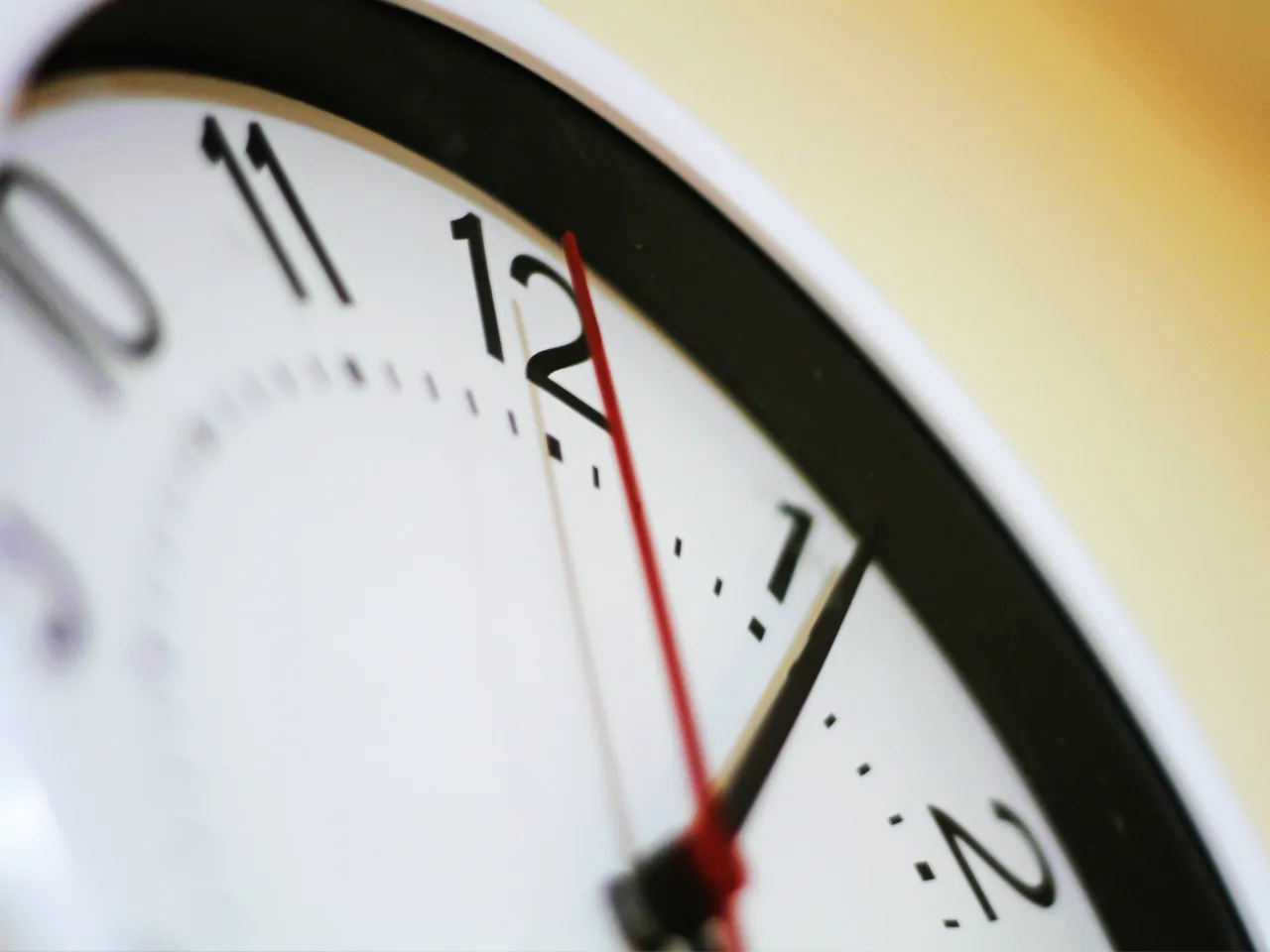 Close-up of a wall clock showing the time as 12:01, with the second hand in red, the minute hand just past 12, and a blurred yellow background.