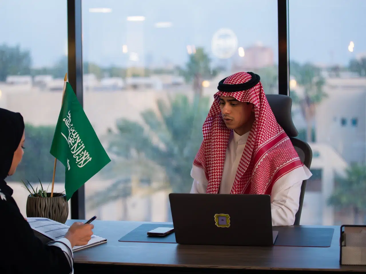 A man in traditional Saudi attire sits at a desk with a laptop, talking to a woman taking notes. A Saudi Arabian flag is displayed on the desk, and a window shows palm trees outside.