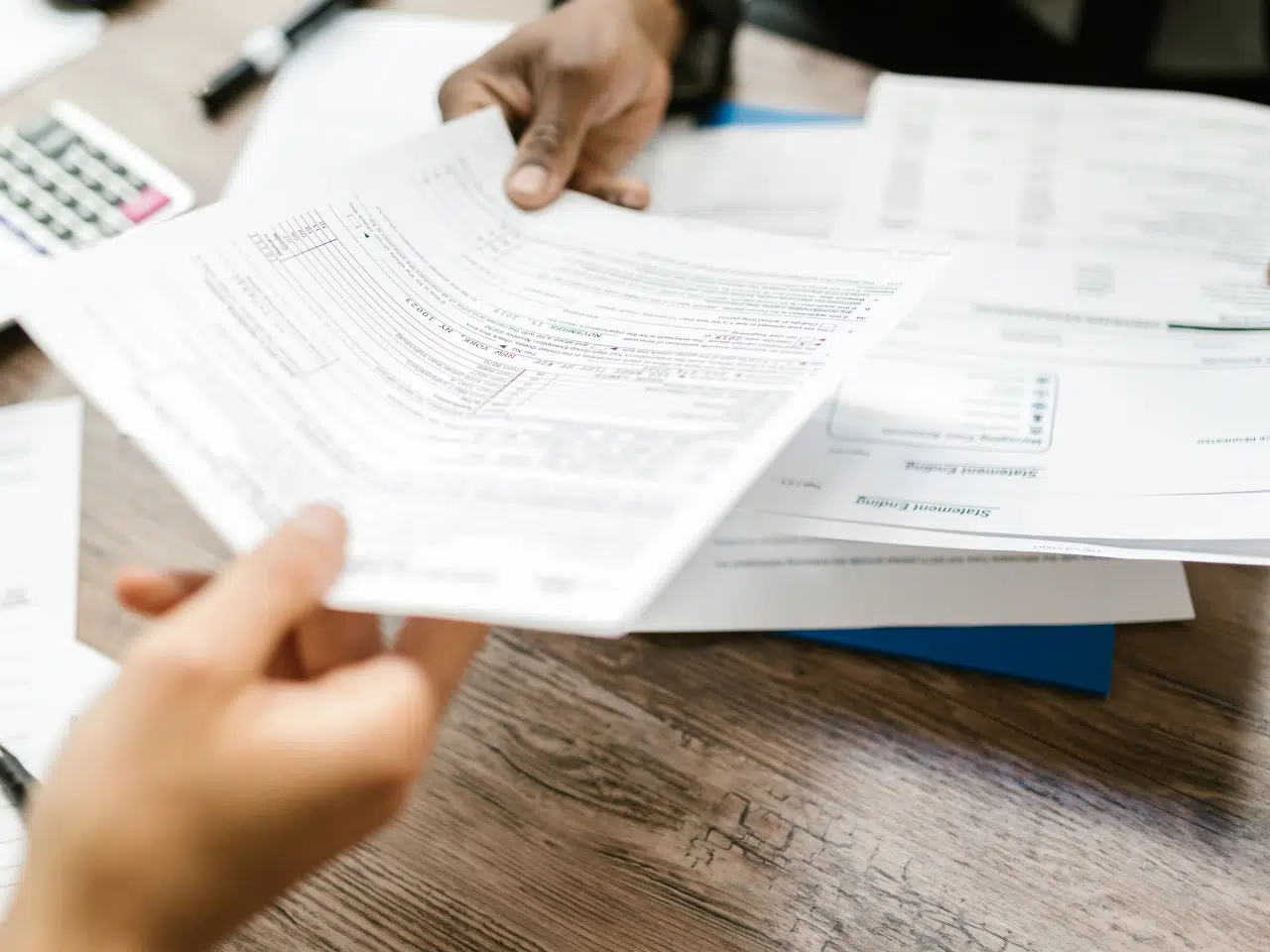 Two people review and exchange paperwork at a table covered with documents, forms, and a calculator, suggesting a financial or administrative setting.