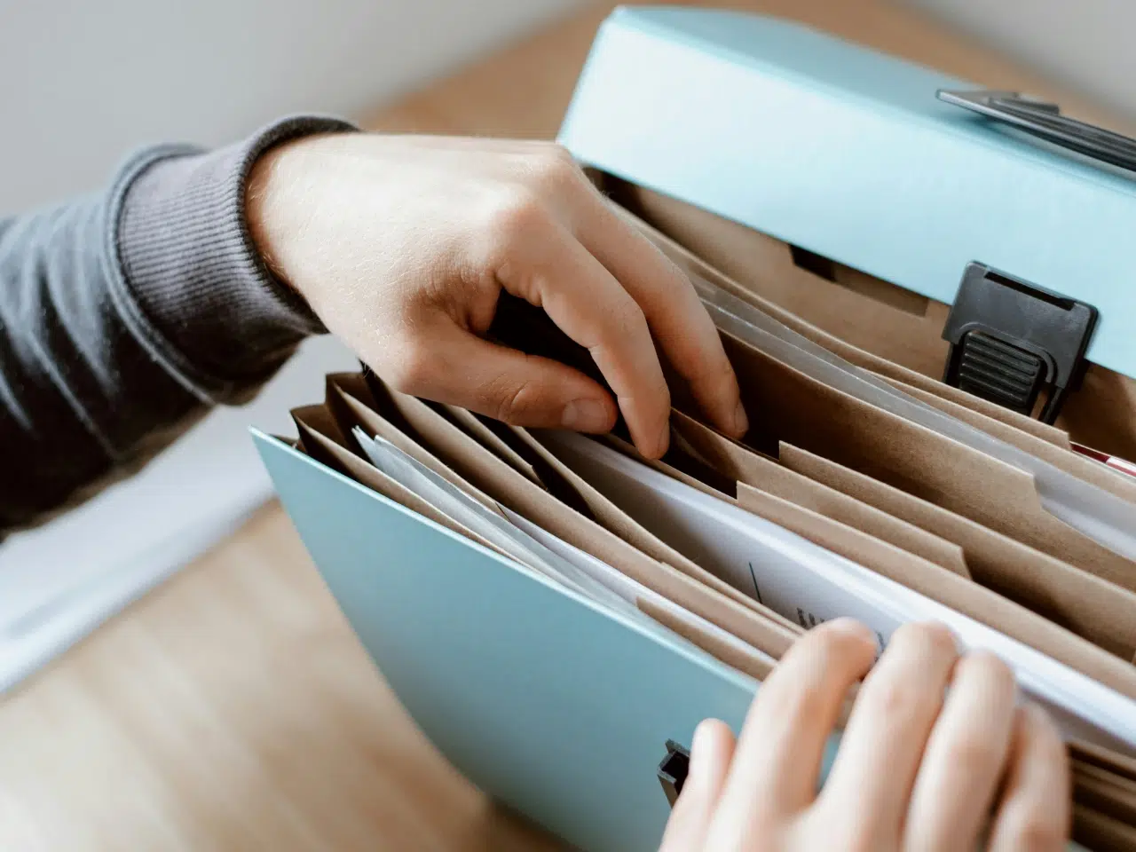 A person’s hands sorting through documents in a blue accordion file folder on a wooden surface.