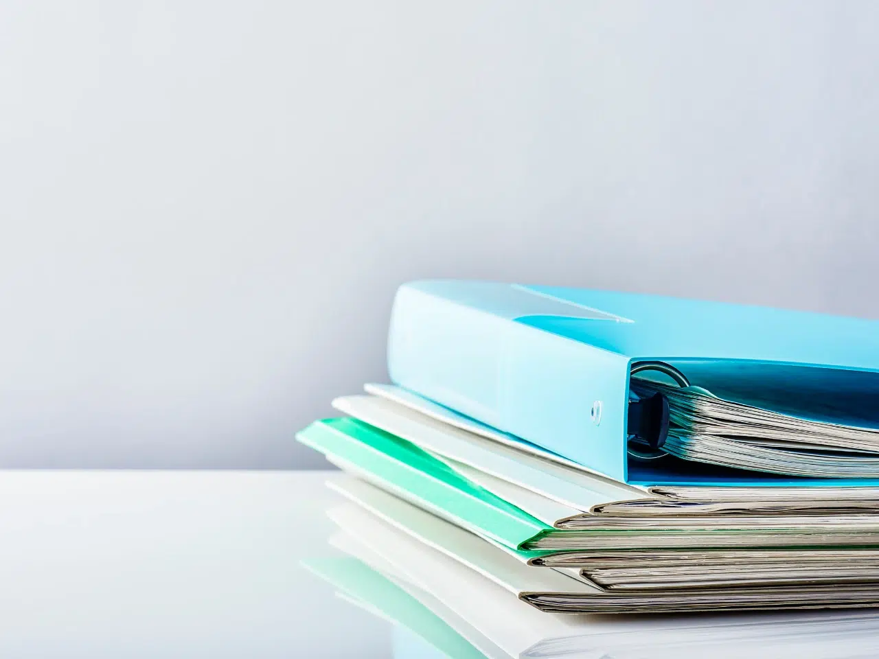 A stack of folders and documents is neatly arranged on a white reflective surface, with a blue binder on top and several files underneath, against a plain light gray background.