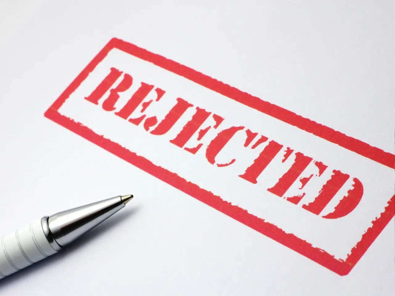 A close-up of a metal pen next to a red REJECTED stamp on white paper.