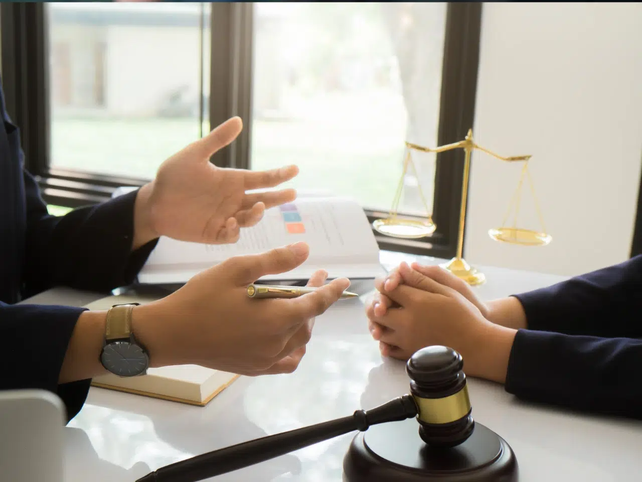 Two people sit at a desk with legal documents; one gestures while holding a pen, the other listens with hands clasped. A gavel and scales of justice are visible on the table, suggesting a legal consultation.