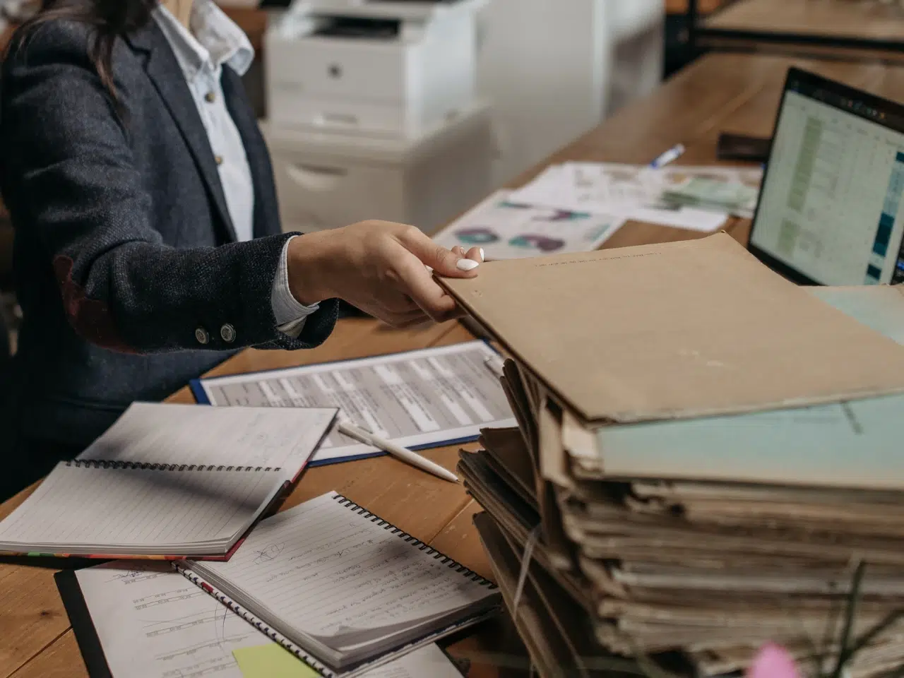 A person in business attire sits at a desk, handing over a stack of large folders. The desk is cluttered with notebooks, documents, and a laptop displaying a spreadsheet.