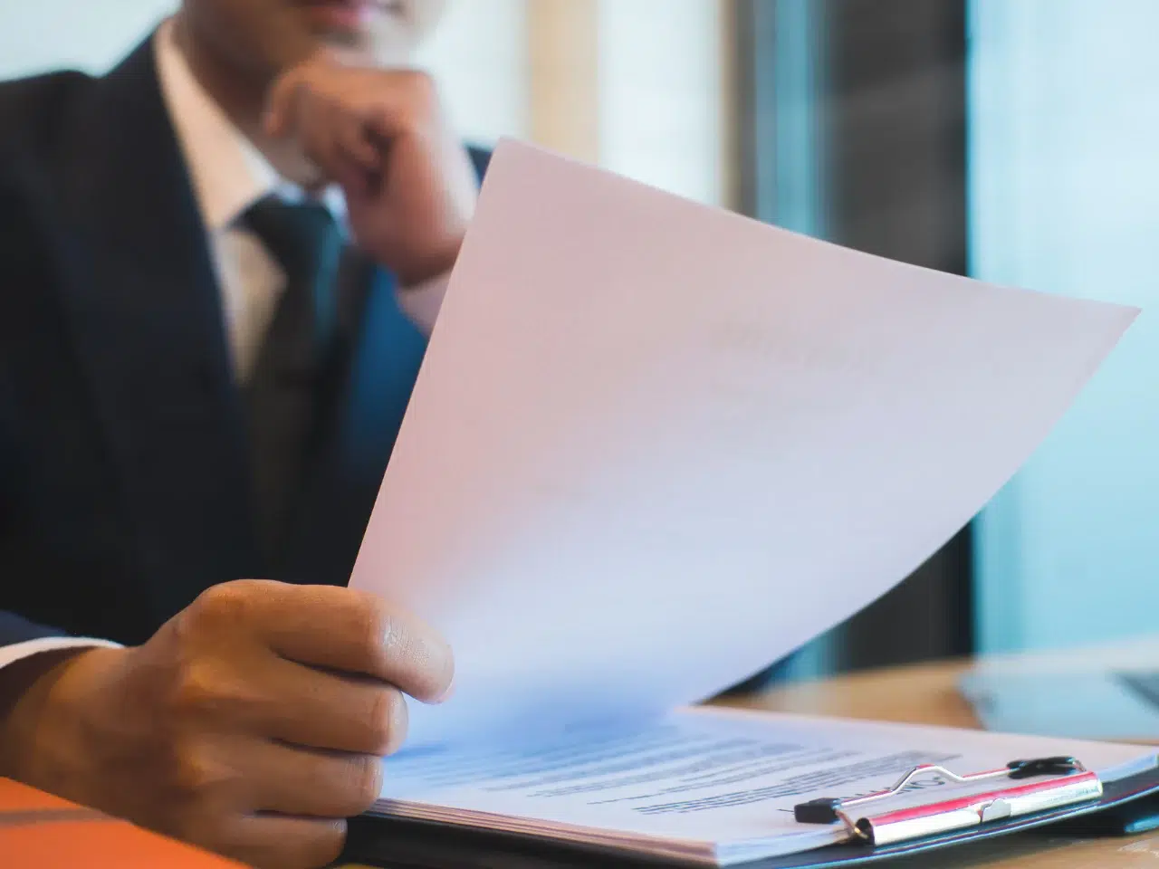A person in a suit sits at a desk, holding and reviewing a stack of papers with one hand, while resting their chin on their other hand in a thoughtful pose. A clipboard and documents are on the table.