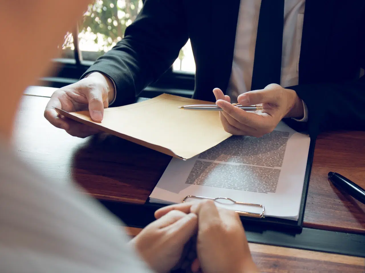 Two people sit at a wooden table; one hands a brown envelope to the other. Documents and a pen are visible on the table, and both individuals appear to be engaged in a discussion.