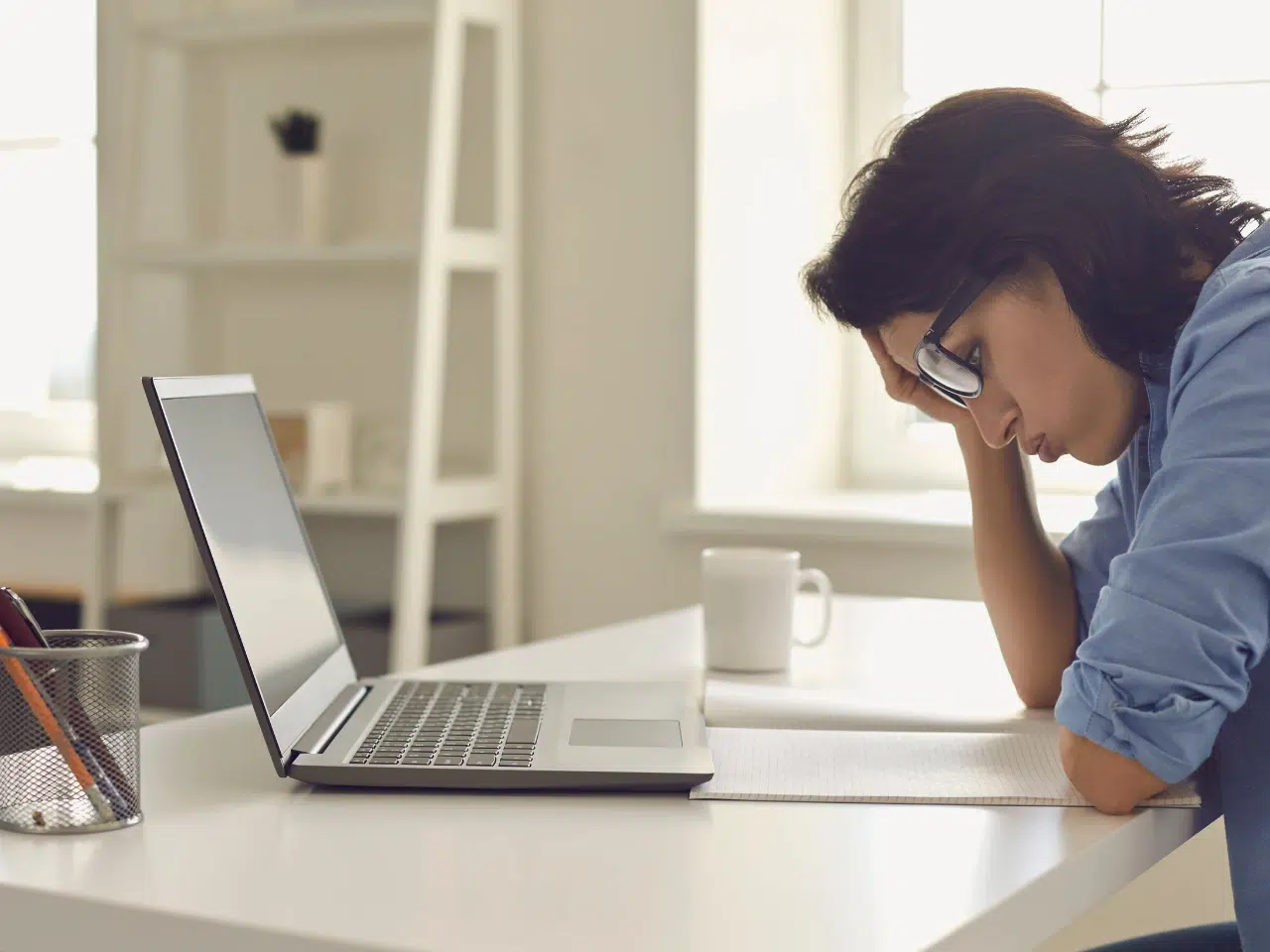 A woman wearing glasses sits at a desk with her head resting on her hand, looking tired or stressed. An open laptop, notebook, and mug are on the desk in front of her.
