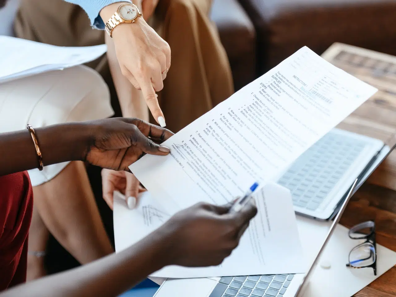 Two people review a printed document together at a desk with an open laptop. One person holds the paper while the other points at a section, suggesting a discussion or analysis of the document’s content.