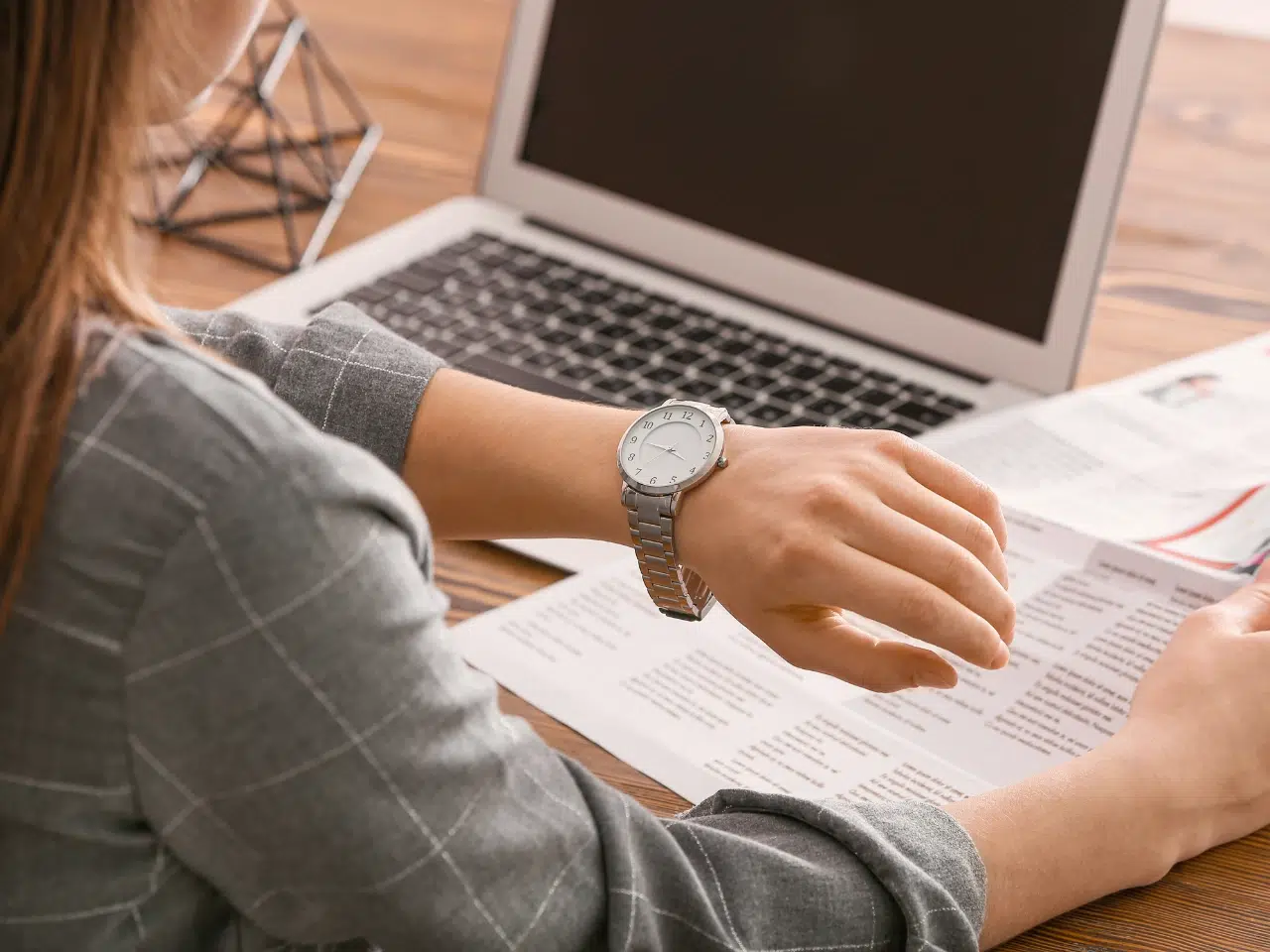 A person in a gray plaid suit looks at their wristwatch while working at a desk with papers and an open laptop.