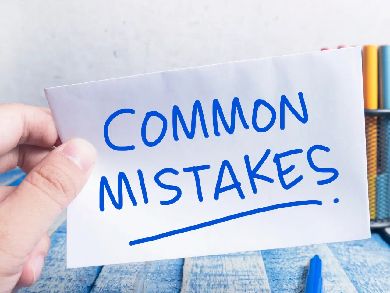 A hand holding a white card with the words COMMON MISTAKES written in bold blue letters. In the background, there are colored pens in a holder on a blue wooden surface.