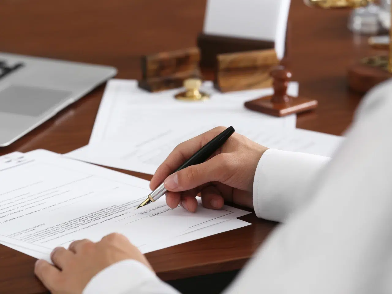 A person in formal attire signs a document at a desk with legal papers, a laptop, and wooden stamps, suggesting a legal or official setting.