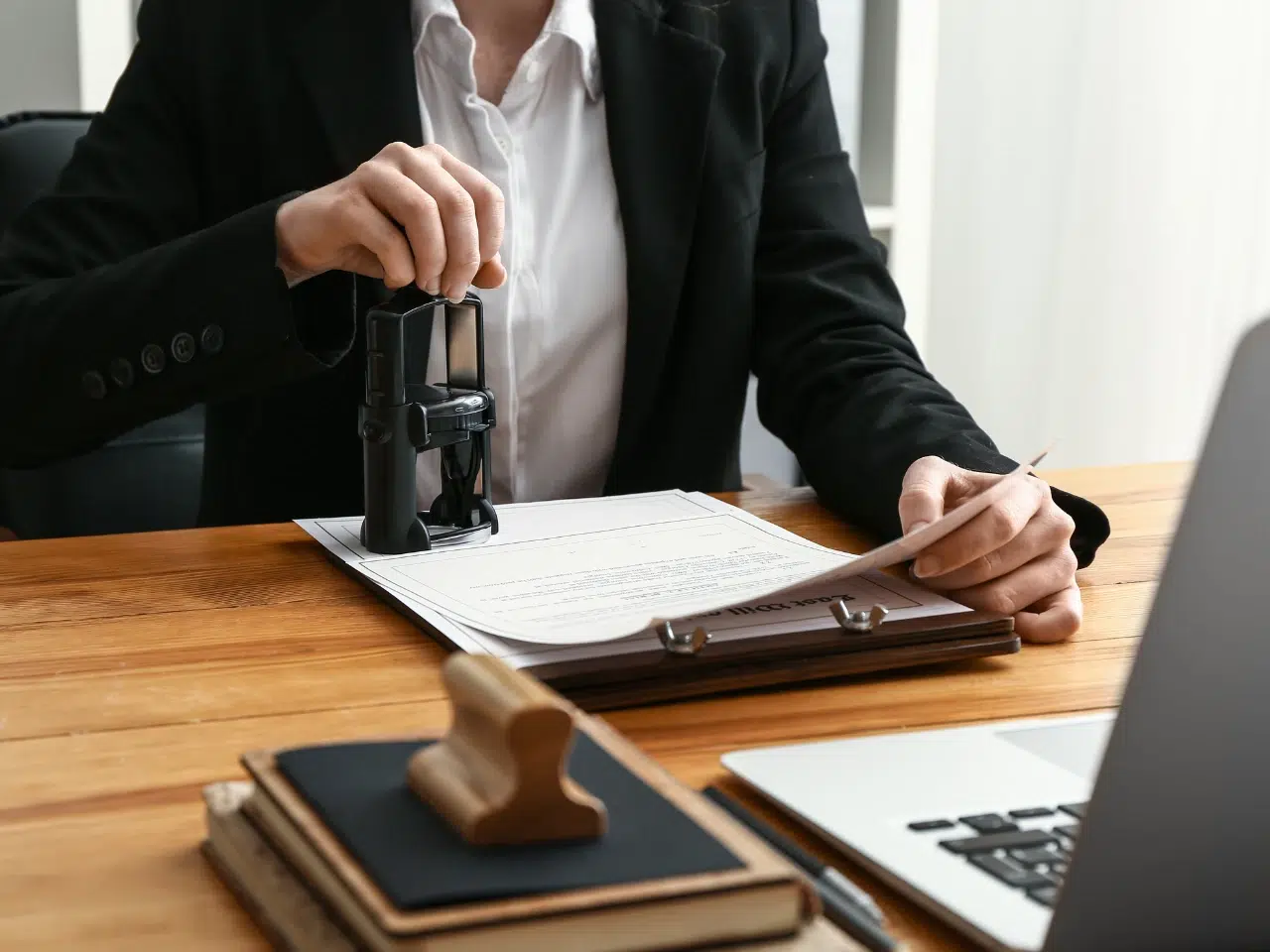 A person in a suit stamps a document on a clipboard at a wooden desk, with a laptop, notebooks, and a wooden stamp visible nearby.
