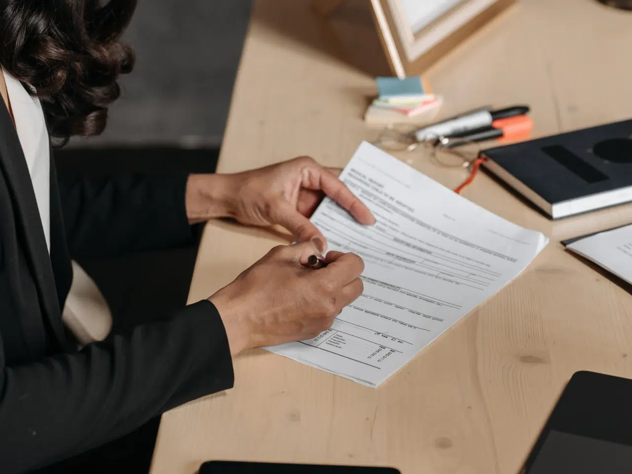 A person in business attire sits at a desk, holding a pen and filling out a form or document. Office supplies, including pens, sticky notes, and a notebook, are visible on the desk.