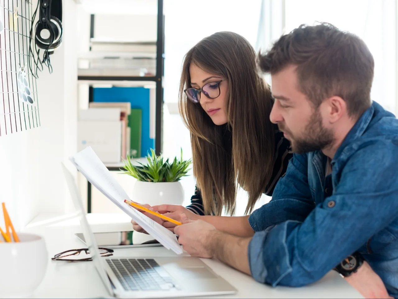 Two people working together at a desk, looking at documents and taking notes next to a laptop and a small potted plant. Both appear focused and are collaborating in a bright office setting.