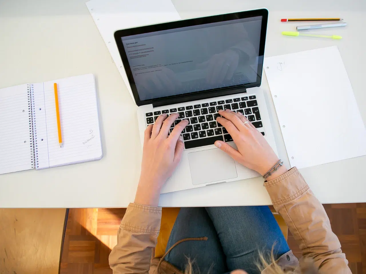Person sitting at a desk typing on a laptop, with a notebook, pencil, and scattered papers nearby. The workspace is organized and well-lit, viewed from above.