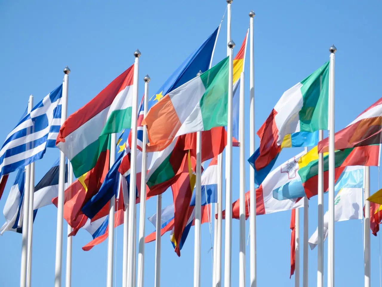 Multiple national flags from different countries wave on flagpoles against a clear blue sky, symbolizing international unity or cooperation.