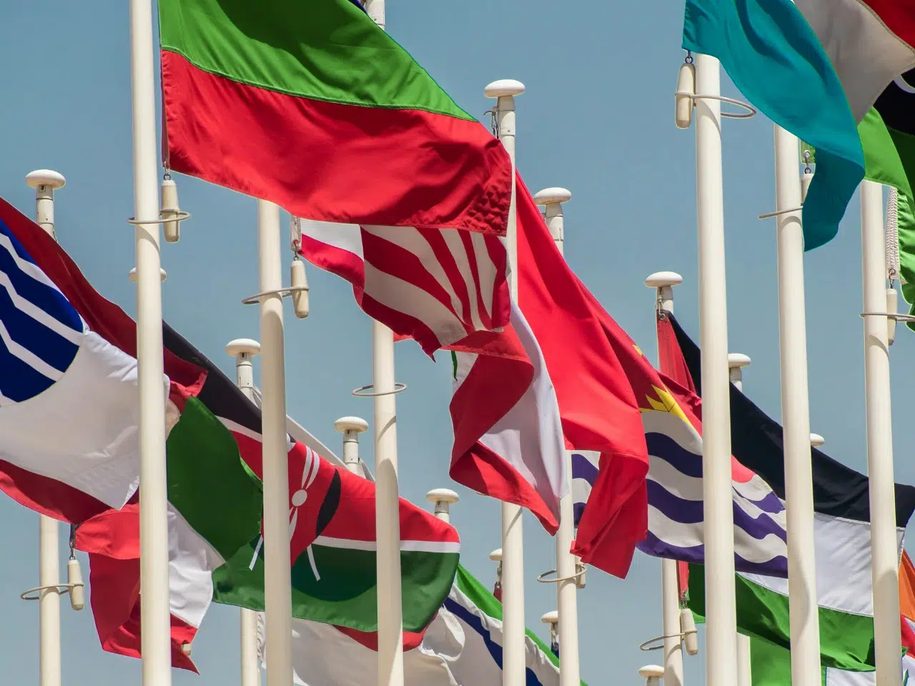 A group of various national flags, each with distinct colors and patterns, waving on tall flagpoles against a clear blue sky.