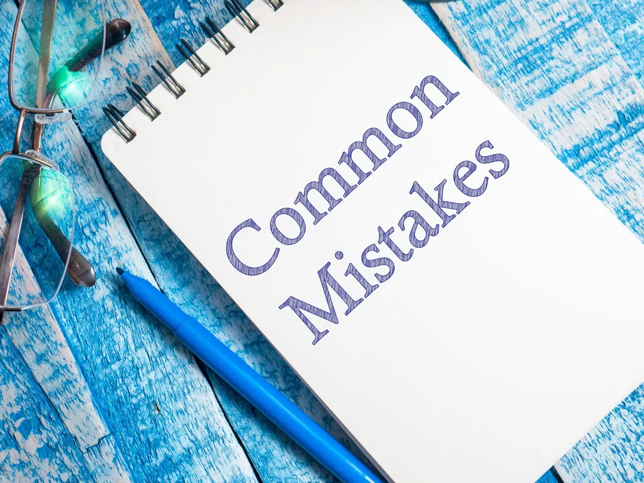 A notepad with “Common Mistakes” written on it, placed on a blue wooden surface next to a blue pen and eyeglasses.