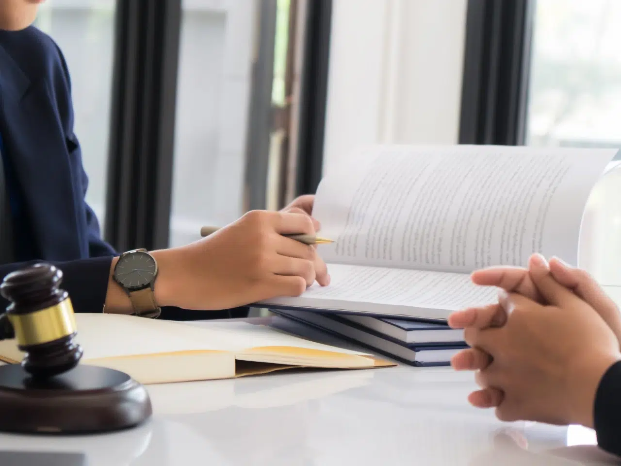 Two people sit across a desk from each other, one holding a pen and reviewing a document, with a stack of books and a judges gavel on the table.