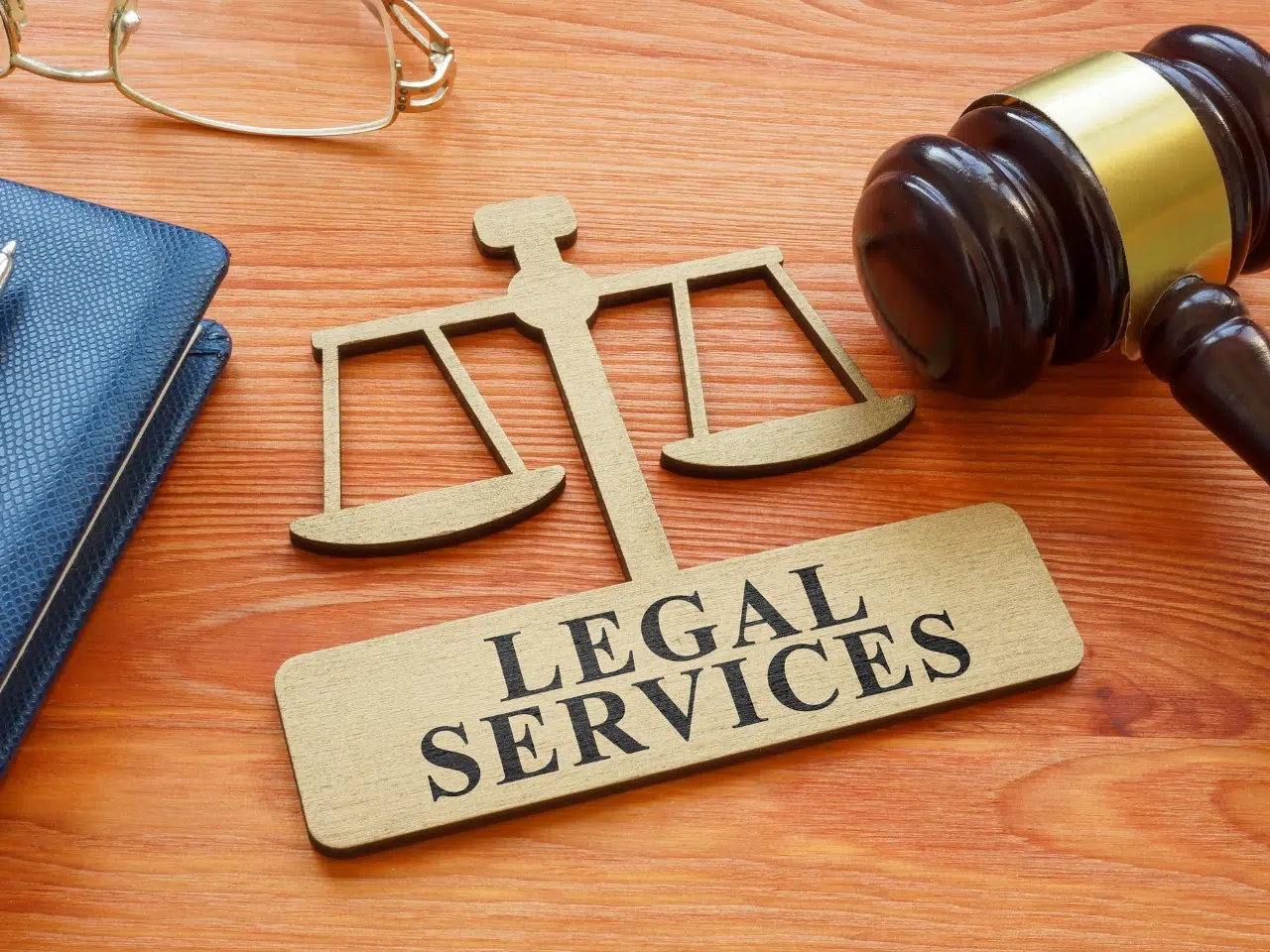 A wooden sign reading Legal Services with a scale of justice symbol, placed on a wooden desk near a gavel, notebook, and eyeglasses.