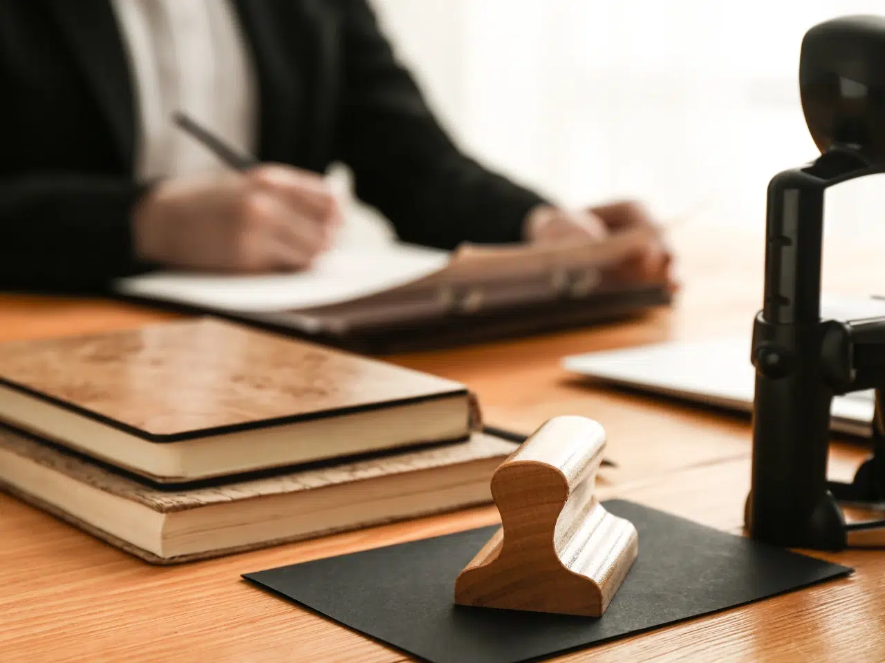 A wooden notary stamp sits on a black envelope in the foreground, with stacked notebooks and a suited person writing at a desk blurred in the background.
