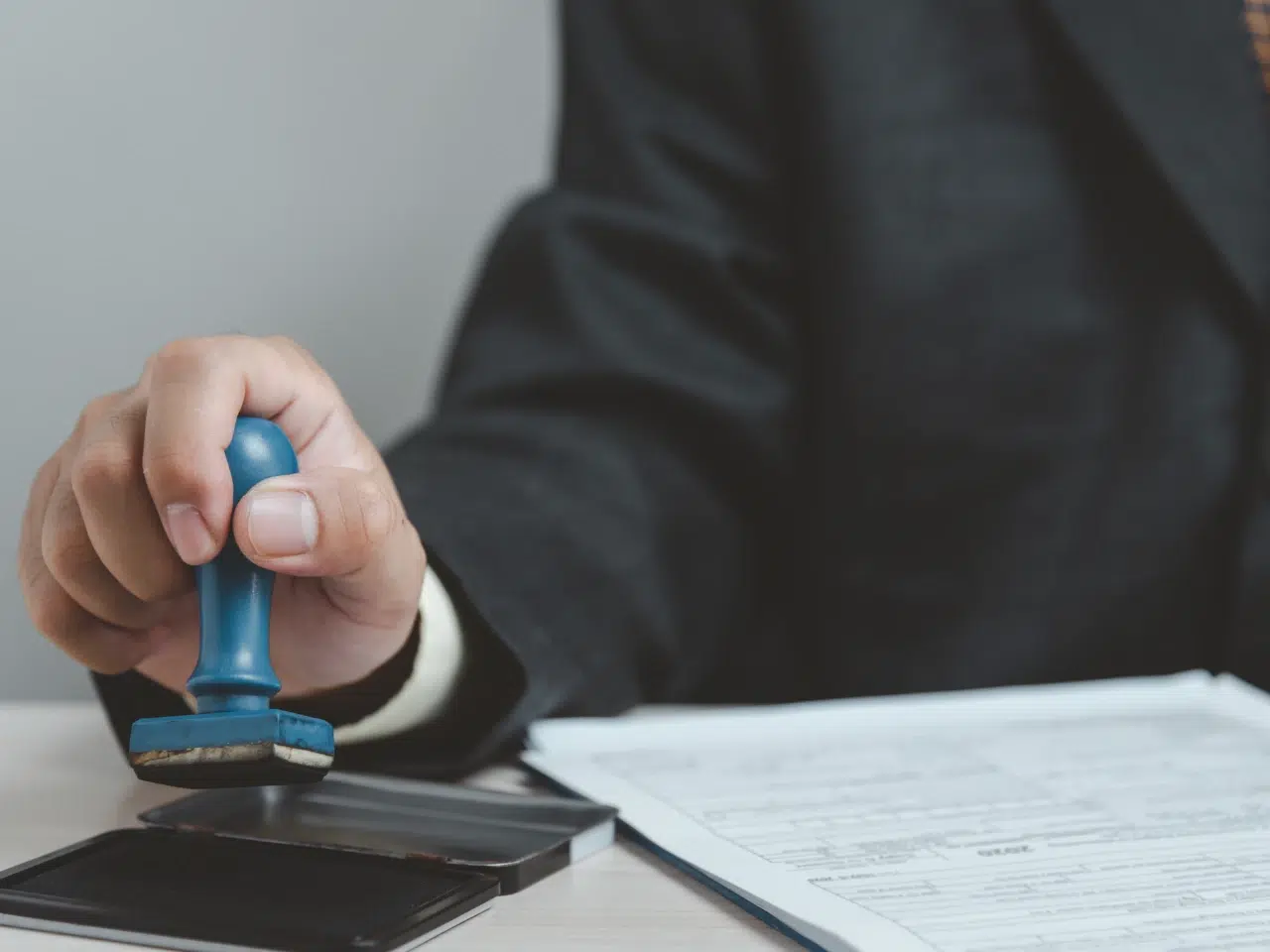 A person in a suit presses a blue rubber stamp into an ink pad beside a stack of paperwork on a desk.