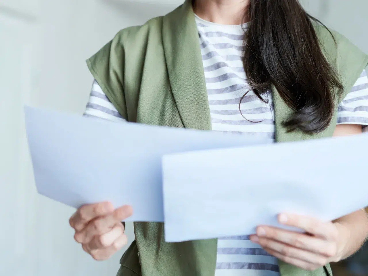 A person wearing a green vest over a gray and white striped shirt is holding and reading two sheets of paper. Their face is not visible in the image.