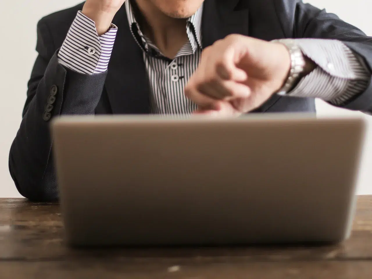 A person in business attire looks at their wristwatch while sitting at a desk with an open laptop, suggesting they are checking the time, possibly feeling rushed or busy.