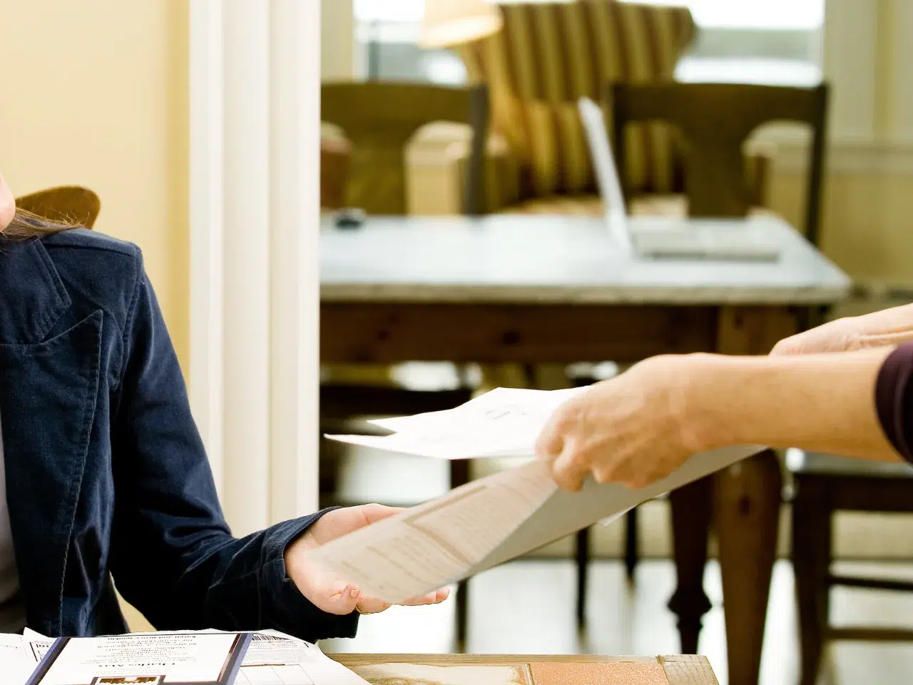 Two people exchanging documents at a table in a sunlit room. Only their hands and arms are visible, with one person handing papers to the other. A desk and chairs are blurred in the background.