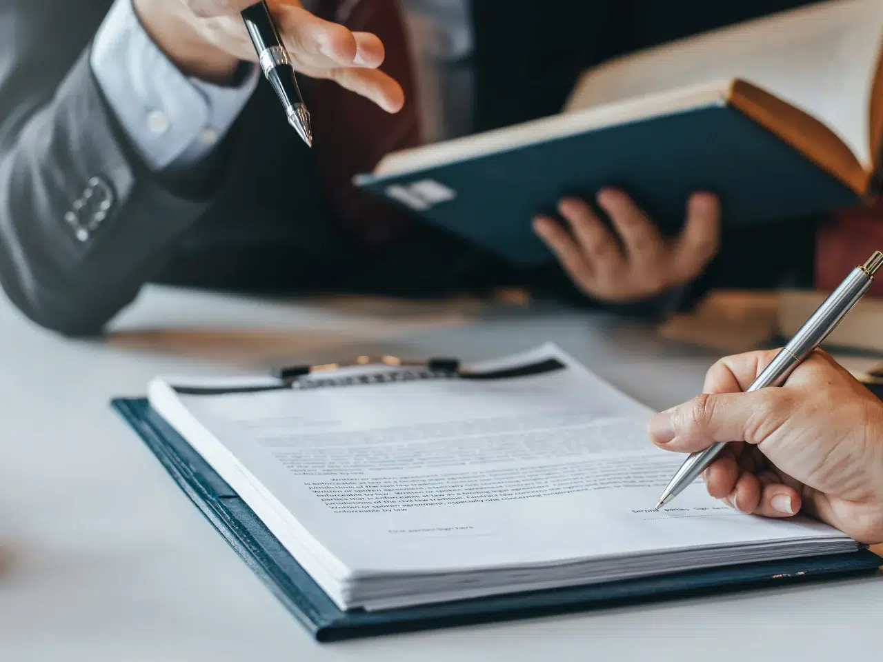 Two people in business attire discuss and review a document on a clipboard; one gestures with a pen while the other prepares to sign the paperwork, with a book open in the background.