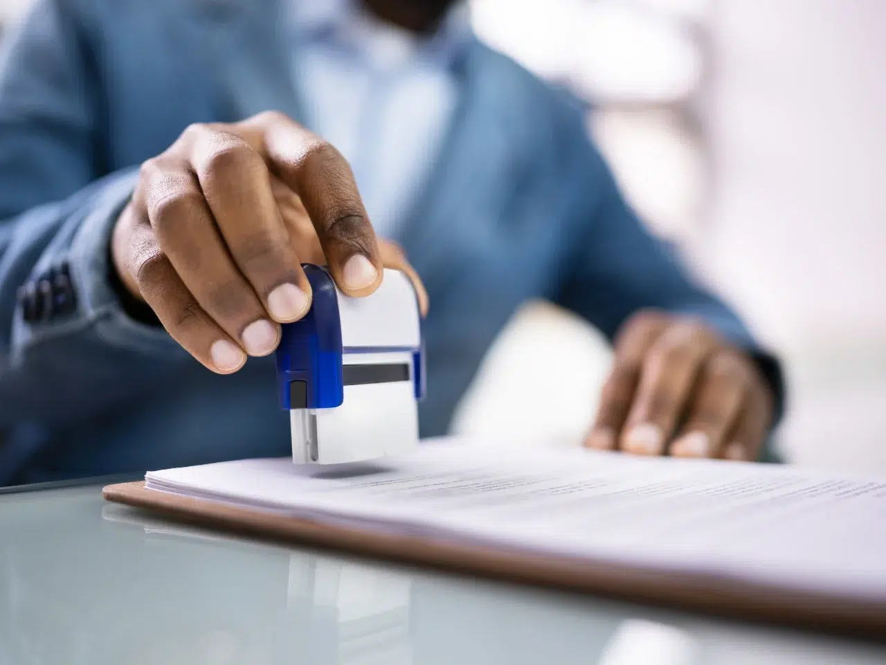 A person in a blue suit presses a blue and white stamp onto a stack of papers on a clipboard, sitting at a glass-top desk. The focus is on the hand and stamp, with text on the papers slightly blurred.