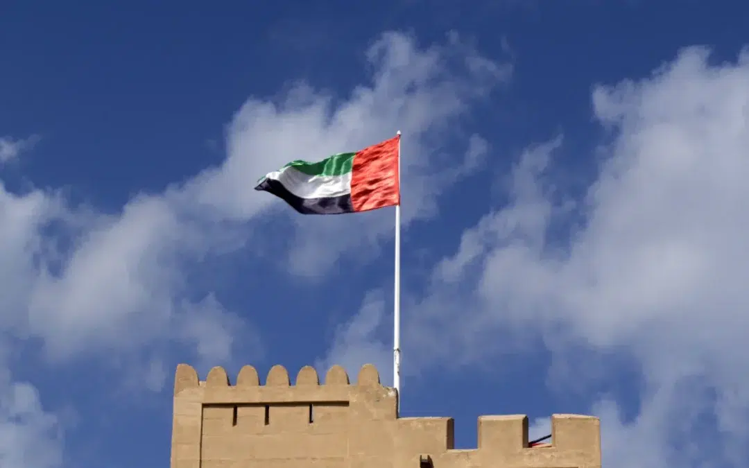 The flag of the United Arab Emirates flies atop a fort-like beige building against a blue sky with scattered white clouds.