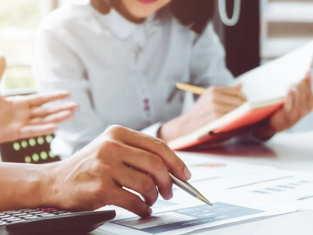 Close-up of people at a desk working together, one person holding a pen and pointing at a document, while another writes in a notebook. Both are dressed in business attire, suggesting a collaborative work environment.