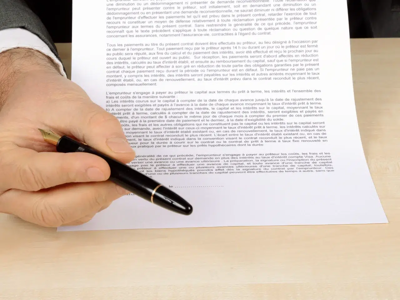 A person holding a black pen is pointing at a printed contract or legal document placed on a light wooden desk. The text on the document is small and densely written.