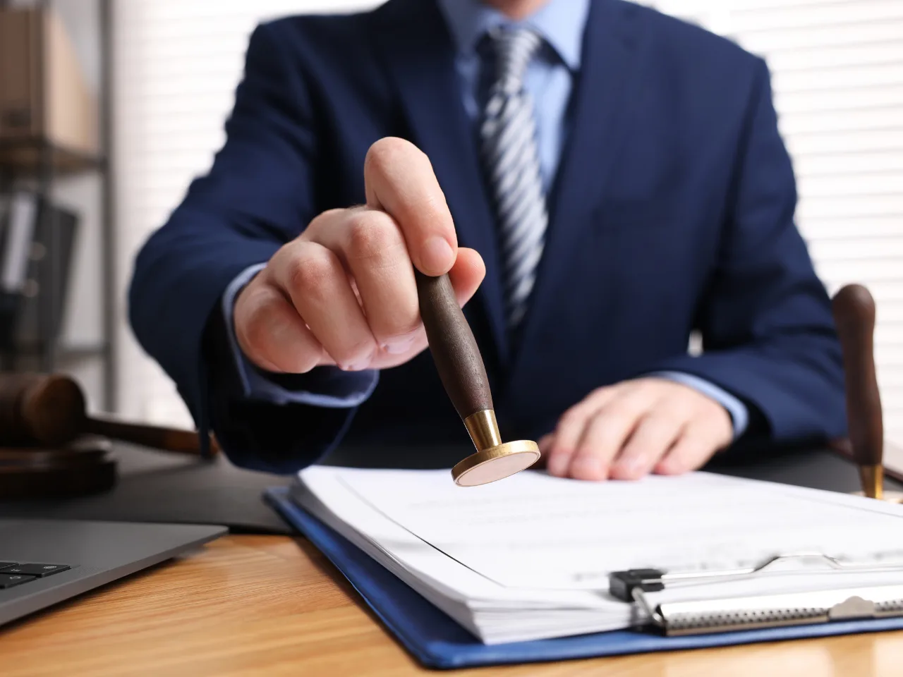 A person in a suit stamps a stack of papers on a desk, with a gavel and clipboard nearby, suggesting an official or legal setting.